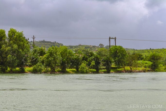Scenic view of riverside cottages surrounded by lush greenery along the shore of Pawna Lake under a cloudy sky.