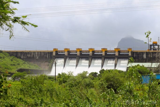 Pawna dam with multiple spillway gates releasing water, surrounded by lush green vegetation and hills in the background under a cloudy sky.