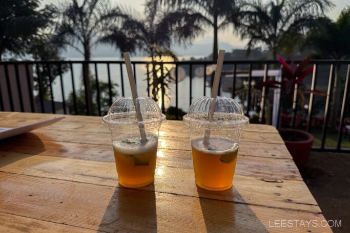 Two cold drinks with lime, set on a wooden table overlooking Pawna Lake at a glamping site, surrounded by palm trees and a scenic view.
