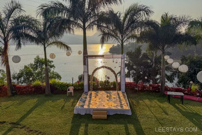 Scenic view of Pawna Lake from a glamping site with a decorated stage, surrounded by palm trees and hanging lanterns, during sunset.