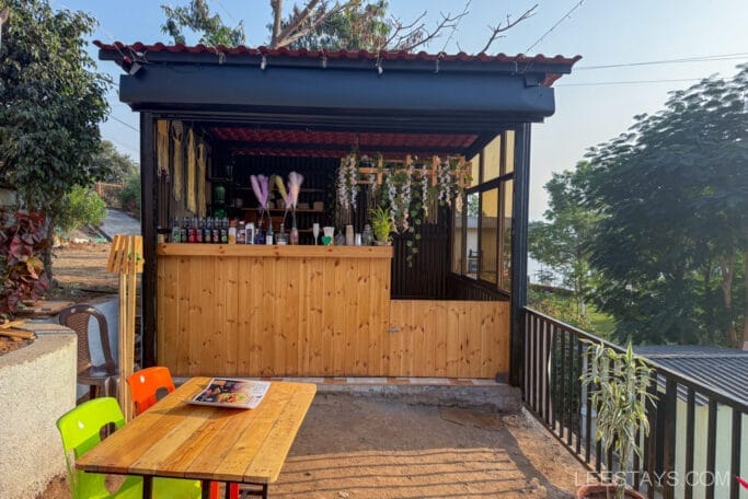 A small wooden outdoor caffee setup with colorful chairs, located near Pawna Lake, ideal for glamping experiences, surrounded by lush greenery.