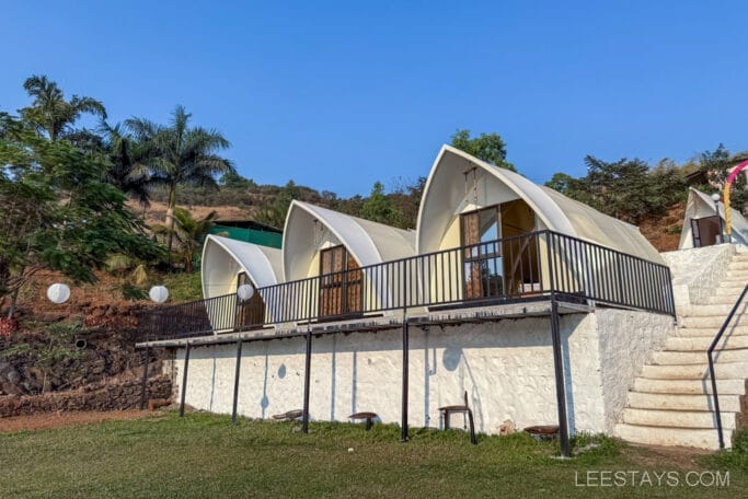 Three modern cabins for glamping at Pawna Lake, featuring unique architectural design with surrounded by greenery and a clear blue sky overhead.