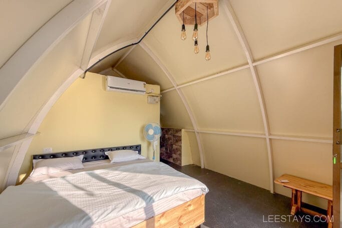 Cozy glamping cabin interior near Pawna Lake, featuring a comfortable bed, ceiling lights, fan, and wooden furnishings, ideal for a camping getaway.