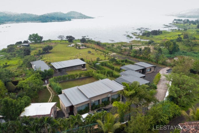 Aerial view of a lakeside resort by Pawna Lake, featuring several modern buildings surrounded by lush greenery and a scenic water body.