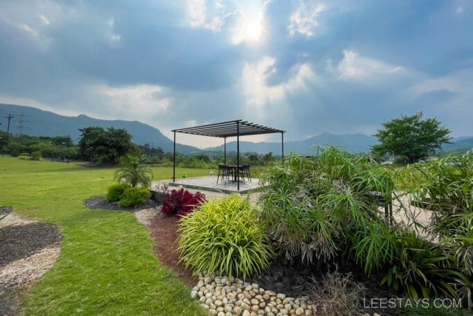 A scenic view of a resort by Pawna Lake, featuring a pergola with chairs set amid lush greenery, with mountains and dramatic clouds in the background.
