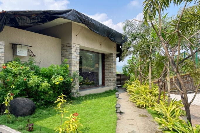 A resort near Pawna Lake featuring a cozy building with stone walls, surrounded by lush greenery and tropical plants under a clear sky.