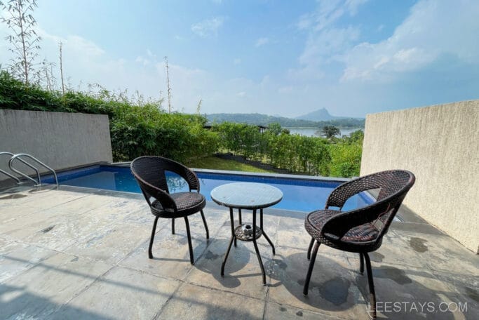 Outdoor seating area by a private pool at a resort near Pawna Lake, with lush greenery and scenic views of hills and the lake in the background under a clear sky.