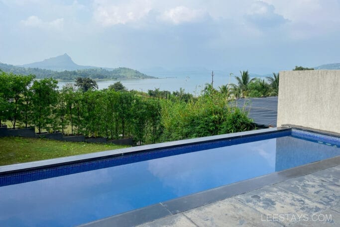 View from a resort overlooking Pawna Lake, featuring a clear swimming pool in the foreground, lush greenery, and distant hills under a blue sky.