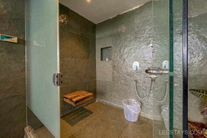 A modern shower with a glass door and stone wall, located in a resort near Pawna Lake, featuring a bench and a clear plastic bucket on the floor.