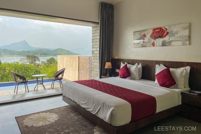 A luxurious resort room overlooking Pawna Lake with a king-sized bed, red cushions, and a view of a patio set by the water and lush green scenery.