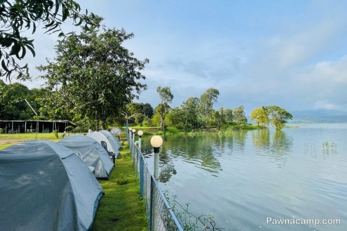 Row of camping tents along the lakeside at Pawna Camp, surrounded by greenery and water.