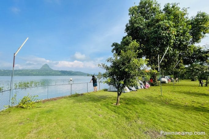 Lakeside camping area with tents and greenery near Pawna Lake, featuring a mountain in the background.