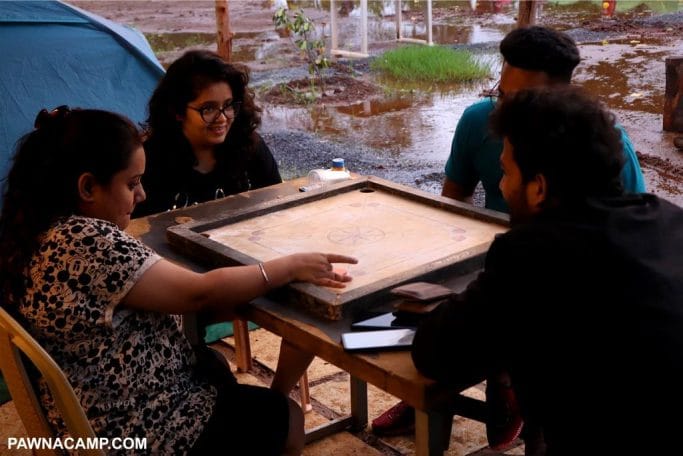 Four individuals engaged in a board game at a camping site with a wet ground and a tent in the background.