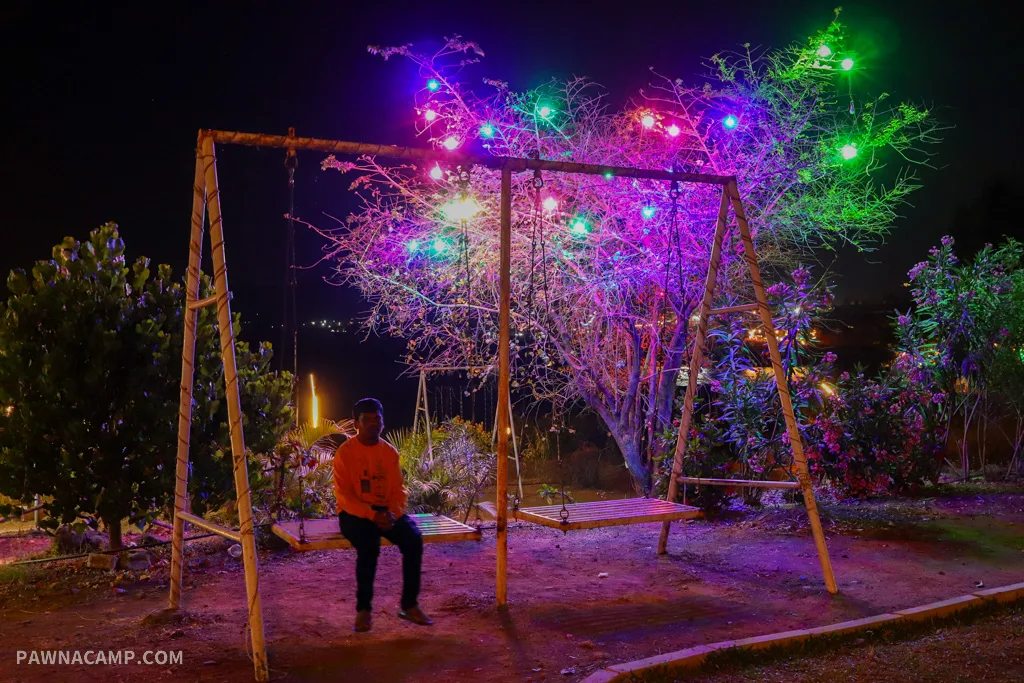 A man sits on a swing set at night with colorful lights in a tree, near Pawna Lake.