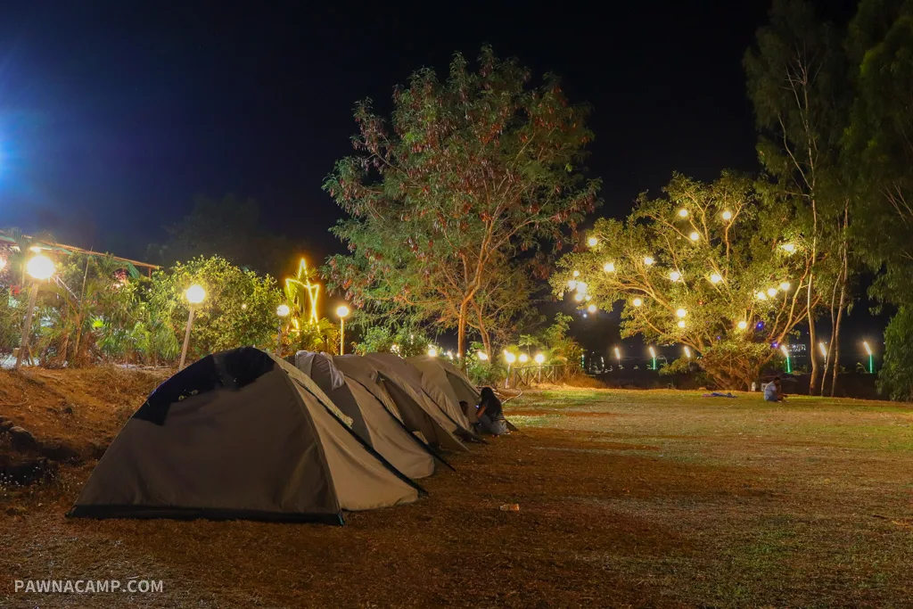 A serene night view of Pawna Lake campsite with lit tents lined up on the grassy ground, surrounded by trees adorned with decorative lights.