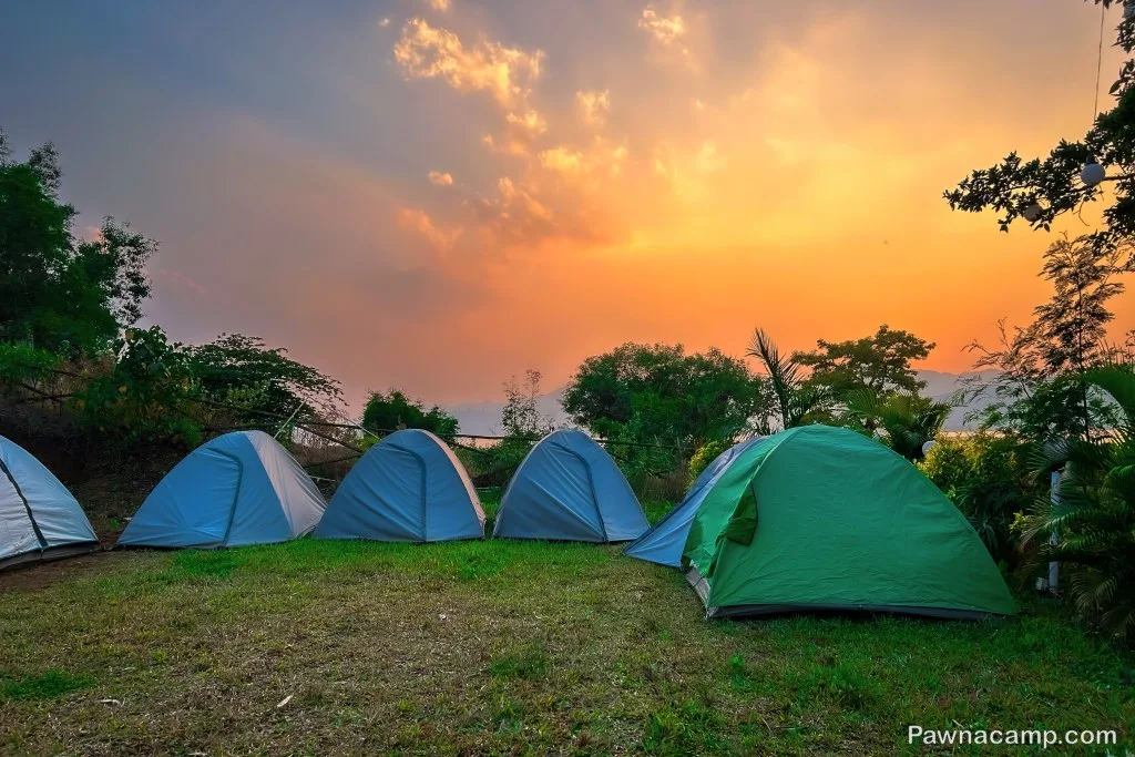 A picturesque view of several tents set up on grassy terrain near Pawna Lake, with a vibrant orange and pink sunset in the sky and trees in the background.