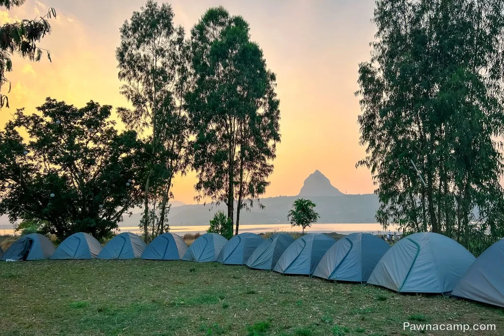 Camping tents set up near Pawna Lake with a serene sunset view, surrounded by lush trees and overlooking a prominent mountain peak in the background.