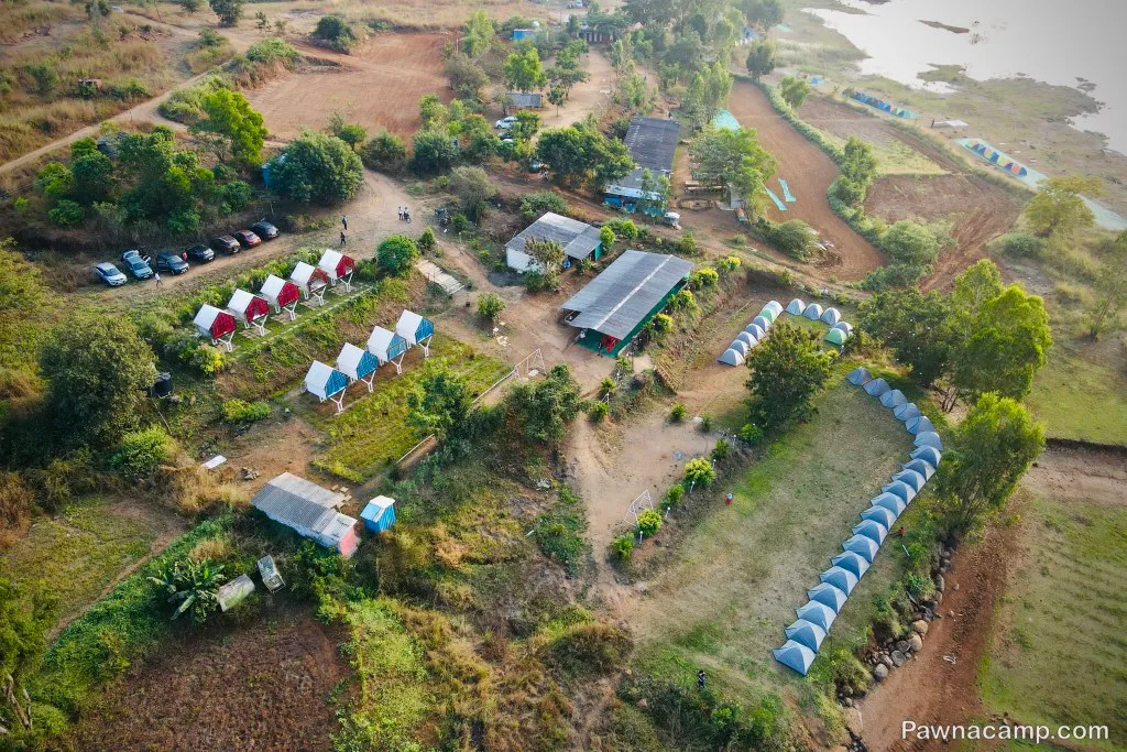 Aerial view of a camping site near Pawna Lake with rows of tents, cabins, trees, and parked cars; the lake is visible in the background.