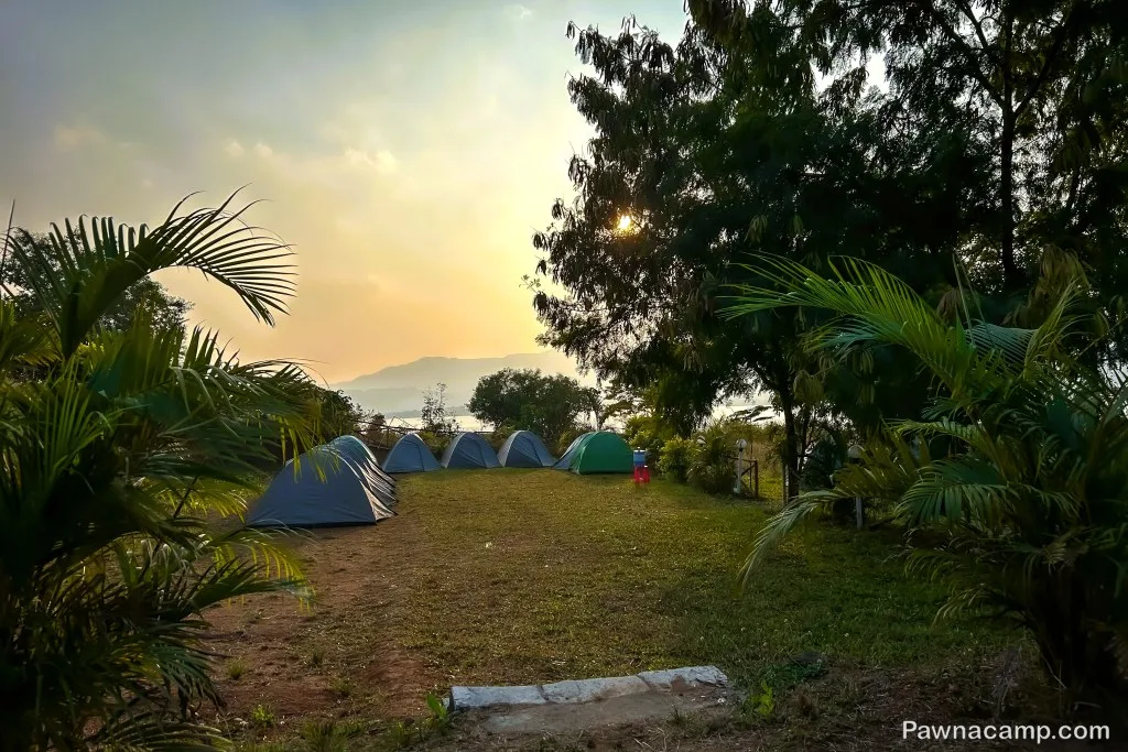 A serene camping site at Pawna Lake, featuring multiple tents set amidst lush greenery and trees, with a tranquil lake and hills in the distant background.