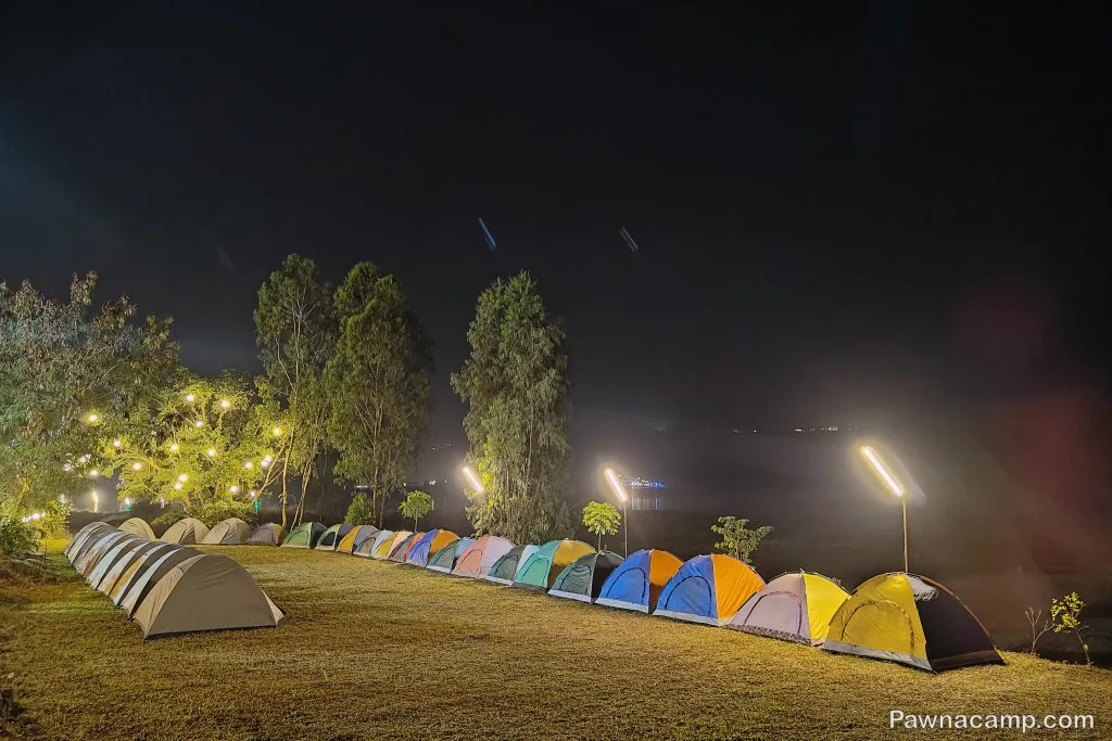 A row of colorful tents set up on a grassy field by Pawna Lake at night, with trees and string lights glowing in the background, creating a serene camping scene.
