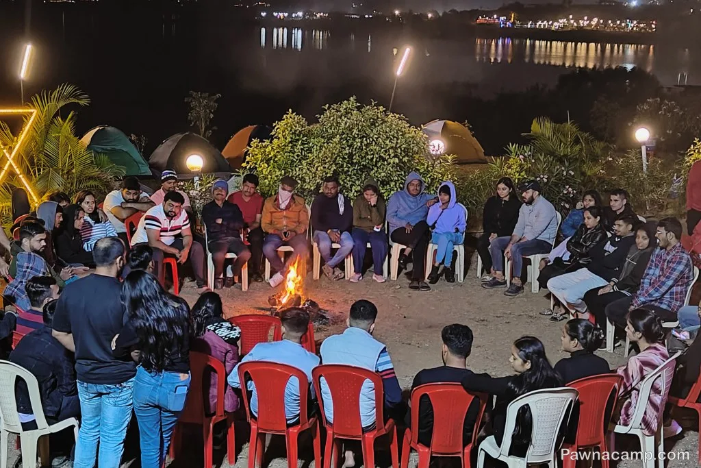 A group of people seated in a circle around a campfire near Pawna Lake at night, with tents in the background and the lake illuminated by distant lights.