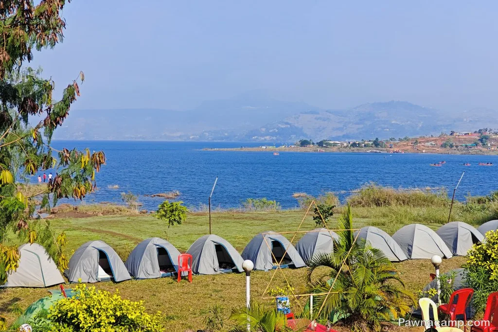 Row of camping tents on the grassy shore of Pawna Lake, with a serene lake view and distant hills under a clear sky.