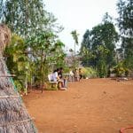 A rustic hut made of straw in a garden setting near Pawna Lake, with people sitting on benches and surrounded by trees.