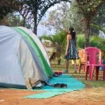 A camping scene near Pawna Lake with a pitched tent, plastic chairs, and a person standing nearby, surrounded by trees and greenery.