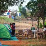 People setting up tents and relaxing at a campsite near Pawna Lake, with trees and huts in the background.