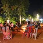 A group of people sitting on chairs around a campfire at night, possibly near Pawna Lake, surrounded by trees and illuminated by string lights.
