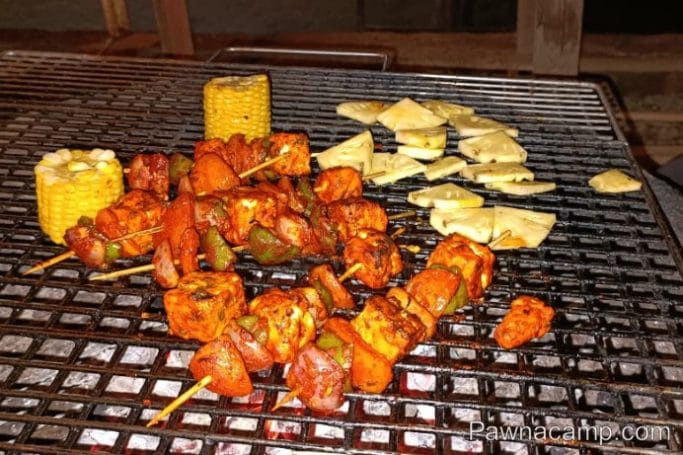 Skewers of marinated vegetables and paneer alongside corn cobs on a barbecue grill.