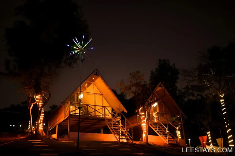 A nighttime view of two illuminated A-frame cottages at Diamond Resort near Pawna Lake, surrounded by trees decorated with fairy lights.
