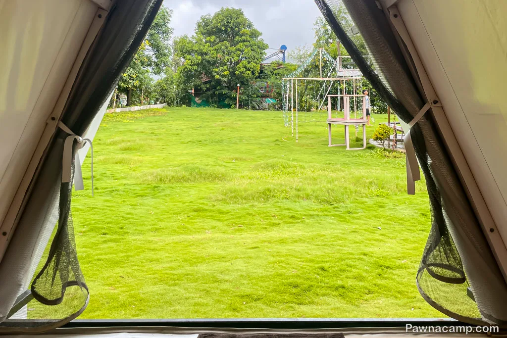 View from inside a tent overlooking a lush green lawn at Pawna Lake, ideal for a peaceful stay, with trees and a play structure visible.