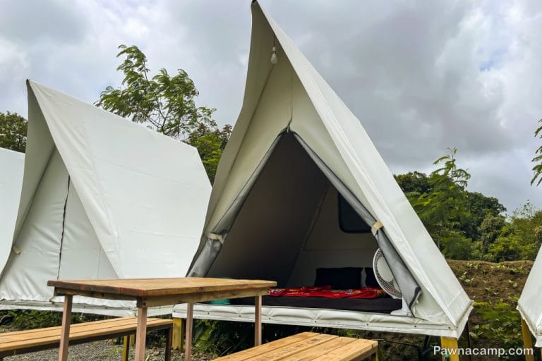 Tents set up for camping near Pawna Lake, featuring a wooden table and benches, surrounded by greenery and cloudy skies.