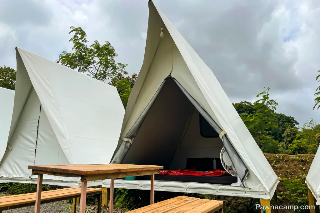 Tents set up for camping near Pawna Lake, featuring a wooden table and benches, surrounded by greenery and cloudy skies.