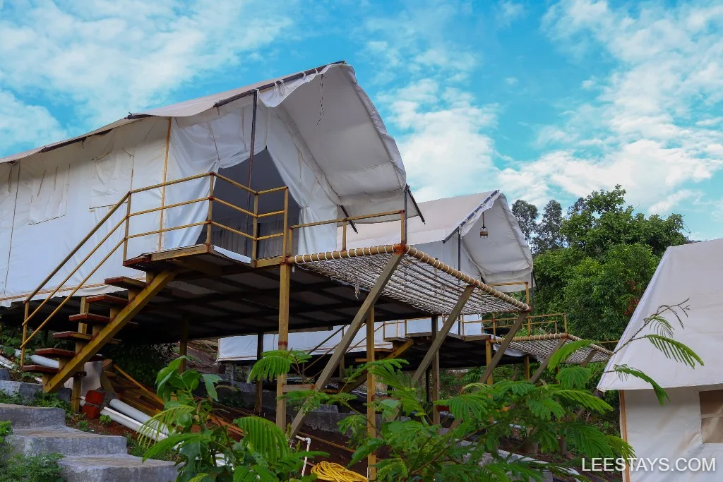 Spacious glamping tents on elevated platforms surrounded by lush greenery near Pawna Lake, set against a bright blue sky.