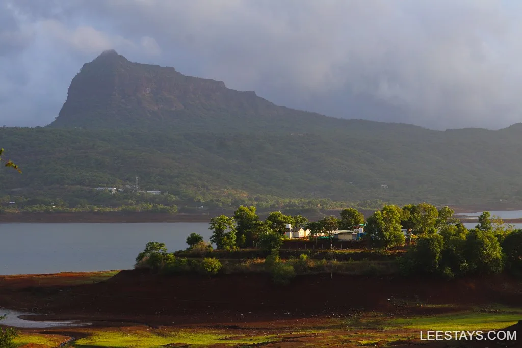 Scenic view of Pawna Lake with lush green hills and a glamping site near the shore, under a cloudy sky.