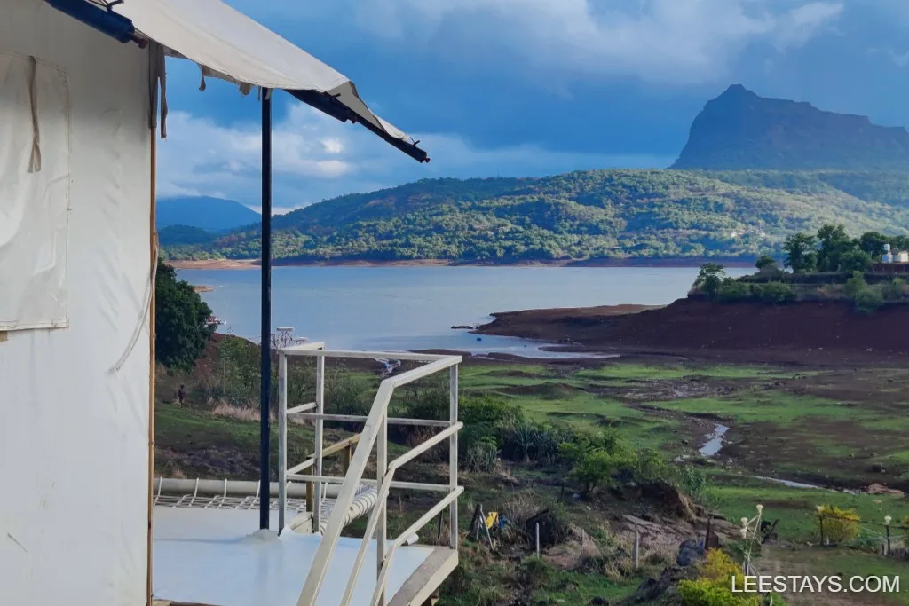 View of Pawna Lake with a glamping tent in the foreground, surrounded by lush green hills and a partly cloudy sky.