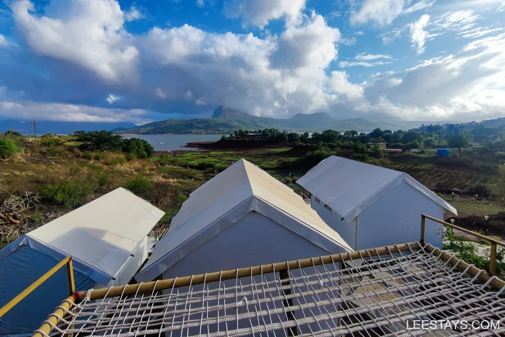 Scenic view of white glamping tents near Pawna Lake with lush greenery and mountains under a partly cloudy sky.