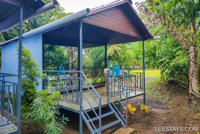 A Pawna cottage with a wooden porch and metal railing, surrounded by lush green foliage.