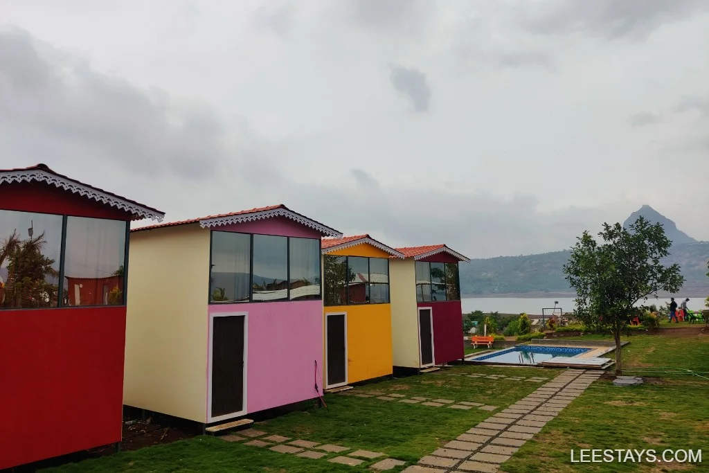 Colorful cottages at a lakeside retreat in Pawna, featuring a pool, lush greenery, and a view of the lake and distant hills under a cloudy sky.