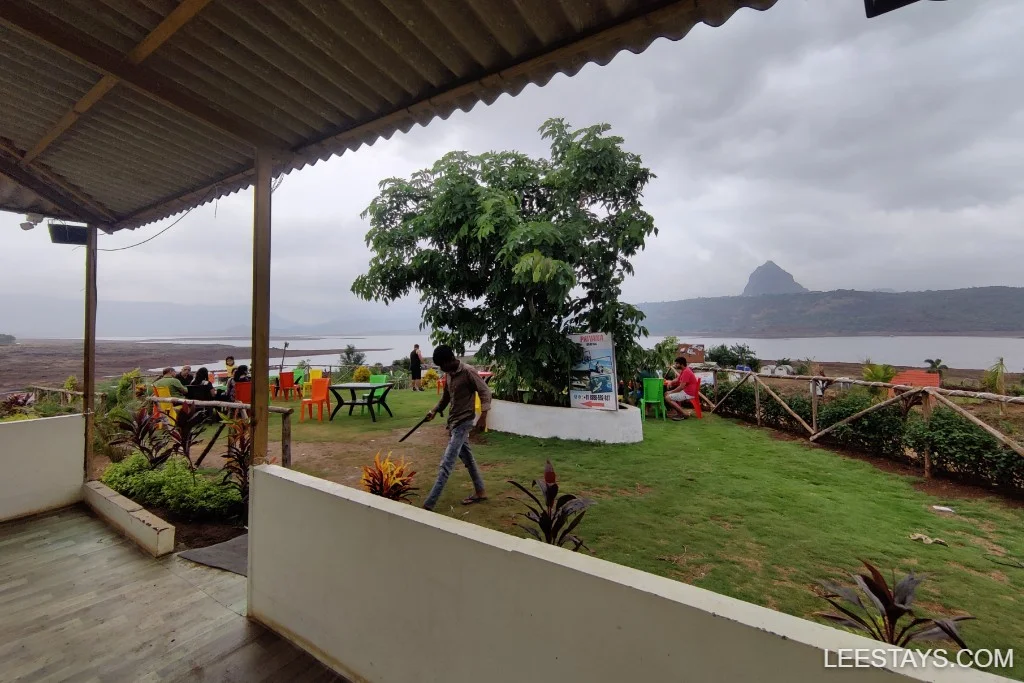 Lakeside retreat in Pawna with people seated at colorful outdoor tables, lush greenery, and a view of the lake under a cloudy sky.