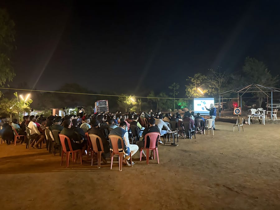 A large group of people seated outdoors at night, facing a person giving a presentation on a screen, with dim lighting and trees in the background.