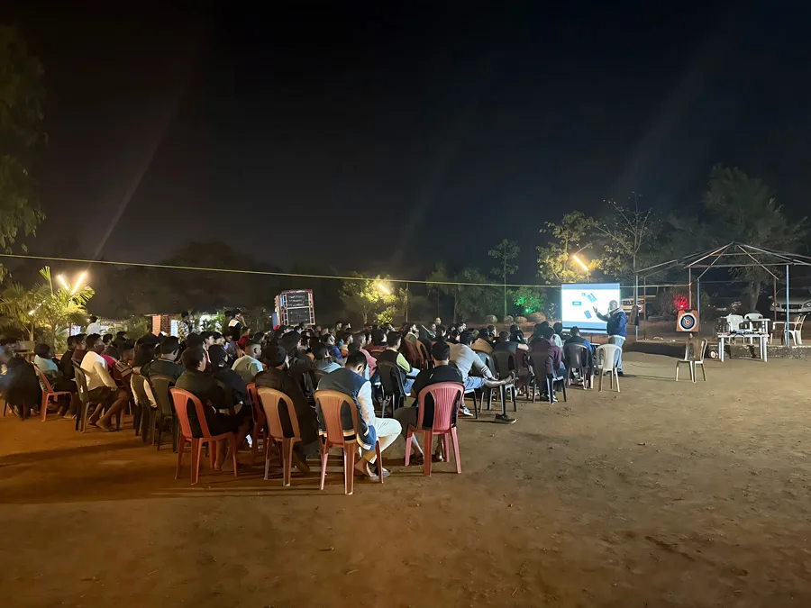 A large group of people seated outdoors at night, facing a person giving a presentation on a screen, with dim lighting and trees in the background.