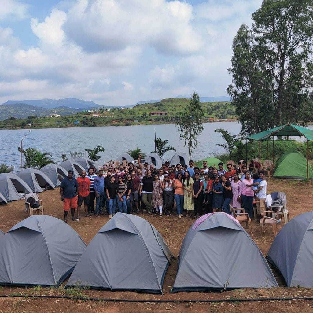 A large group of people gathered at a lakeside campsite with numerous tents, surrounded by greenery and mountains under a cloudy sky.