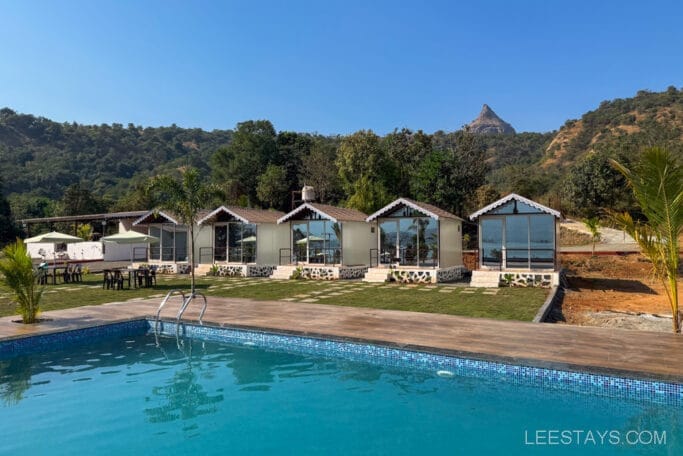 Budget stay near Pawna Lake with pool, featuring multiple small cabins, surrounded by greenery and hills under a clear blue sky.