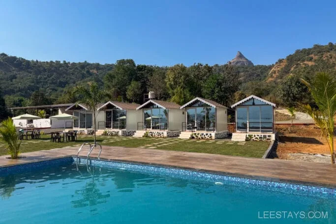 Budget stay near Pawna Lake with pool, featuring multiple small cabins, surrounded by greenery and hills under a clear blue sky.