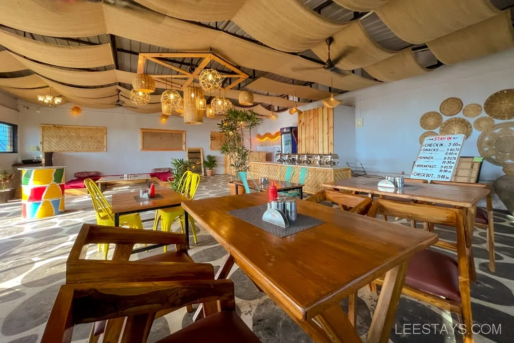 Interior of a restaurant near glass rooms at Pawna Lake, featuring wooden tables, colorful chairs, woven lights, and artistic decor.