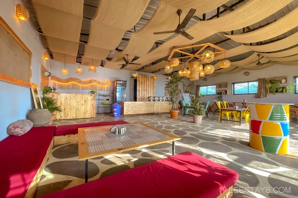 Interior of a glass room near Pawna Lake, featuring modern decor with hanging lights, colorful seating, and large windows letting in natural light.