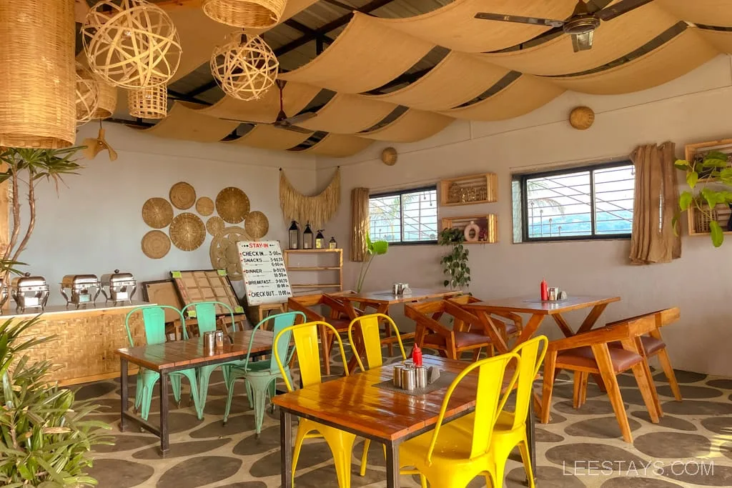 A cozy dining area with wooden tables and colorful chairs in glass rooms near Pawna Lake, featuring ceiling decor and rustic accents.